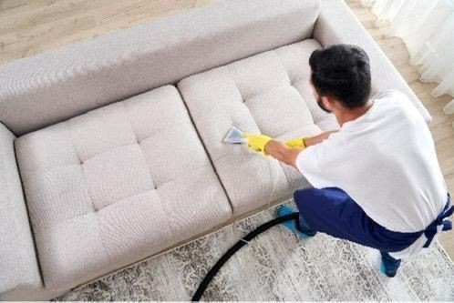 Person wearing gloves using a vacuum cleaner to clean a light-colored sofa in a living room.