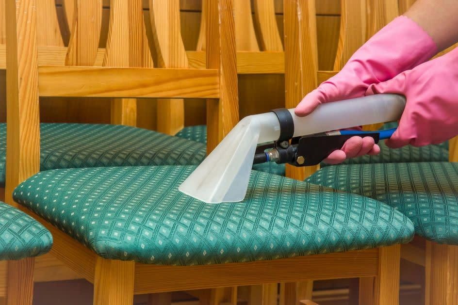 Person cleaning a green upholstered chair with a vacuum tool, wearing pink rubber gloves.