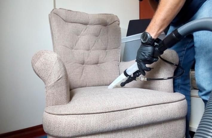 Person cleaning a beige fabric armchair with a vacuum cleaner, wearing a black glove and watch.